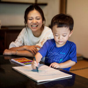 Smiling Japanese woman and little boy sitting at a table, drawing on white paper with colouring pens.