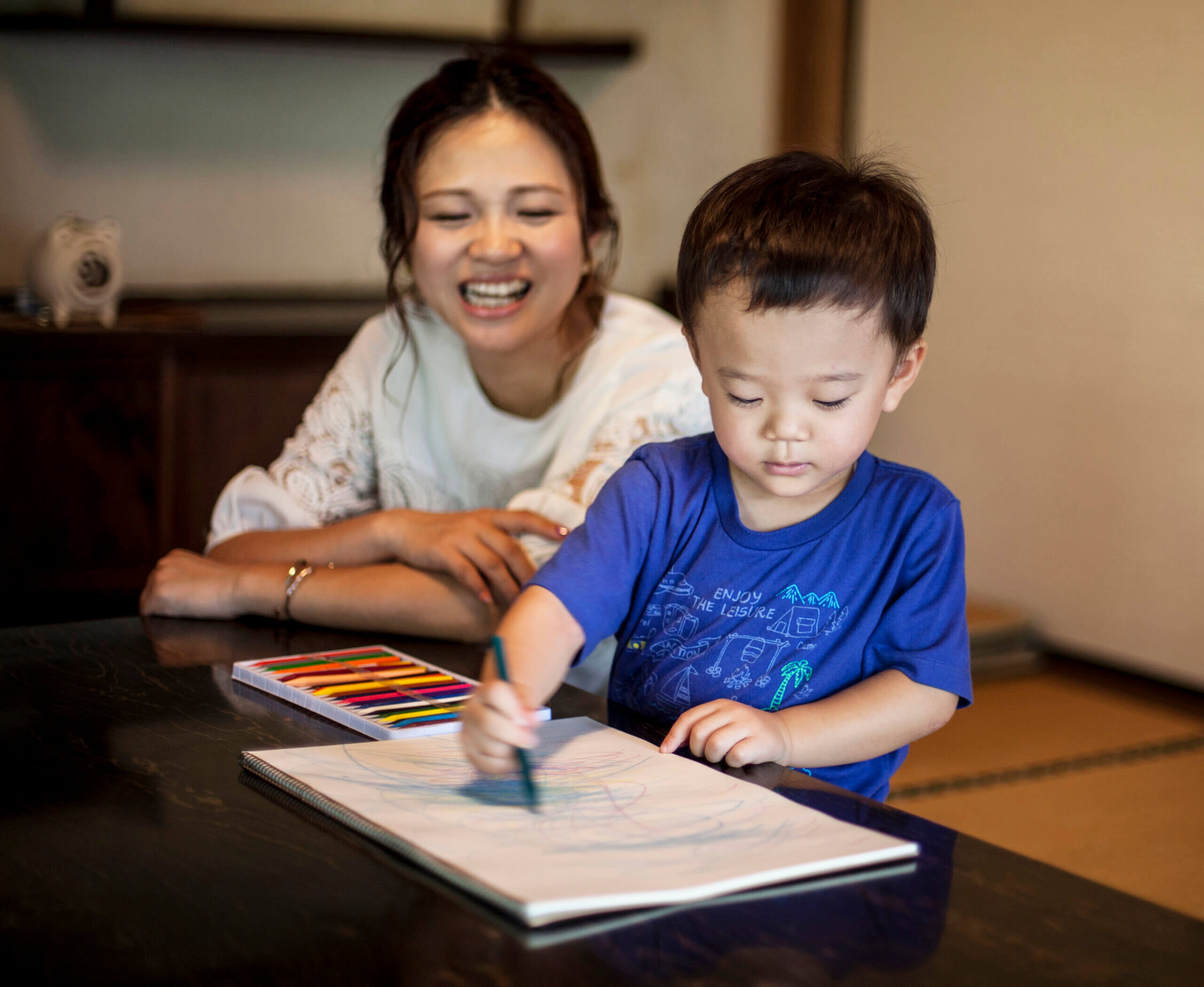Smiling Japanese woman and little boy sitting at a table, drawing on white paper with colouring pens.